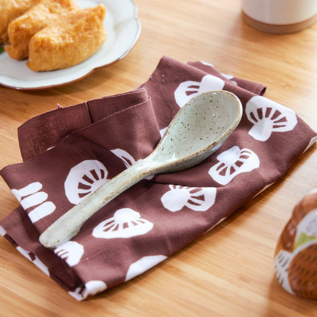 Japanese tenugui cloth with white mushroom patterns on brown fabric, folded neatly beneath a ceramic ramen spoon.