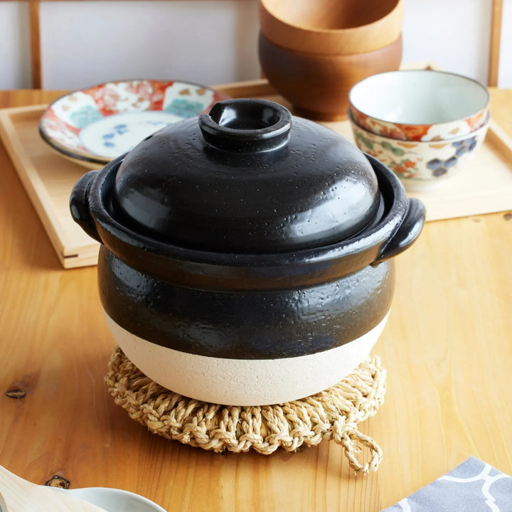 Traditional Japanese black and white donabe clay pot with lid, set on a woven trivet for authentic home-cooked dining.