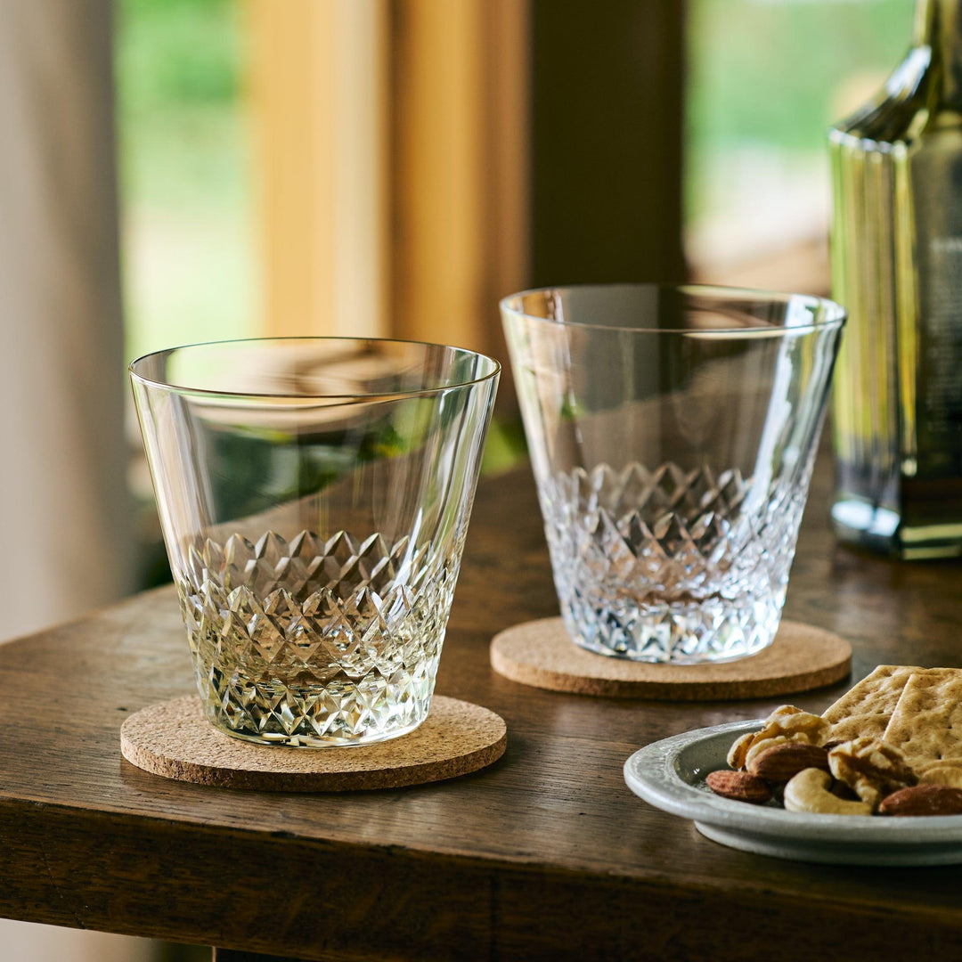 Two empty diamond-cut Japanese whiskey glasses on cork coasters, with snacks and a bottle in the background.