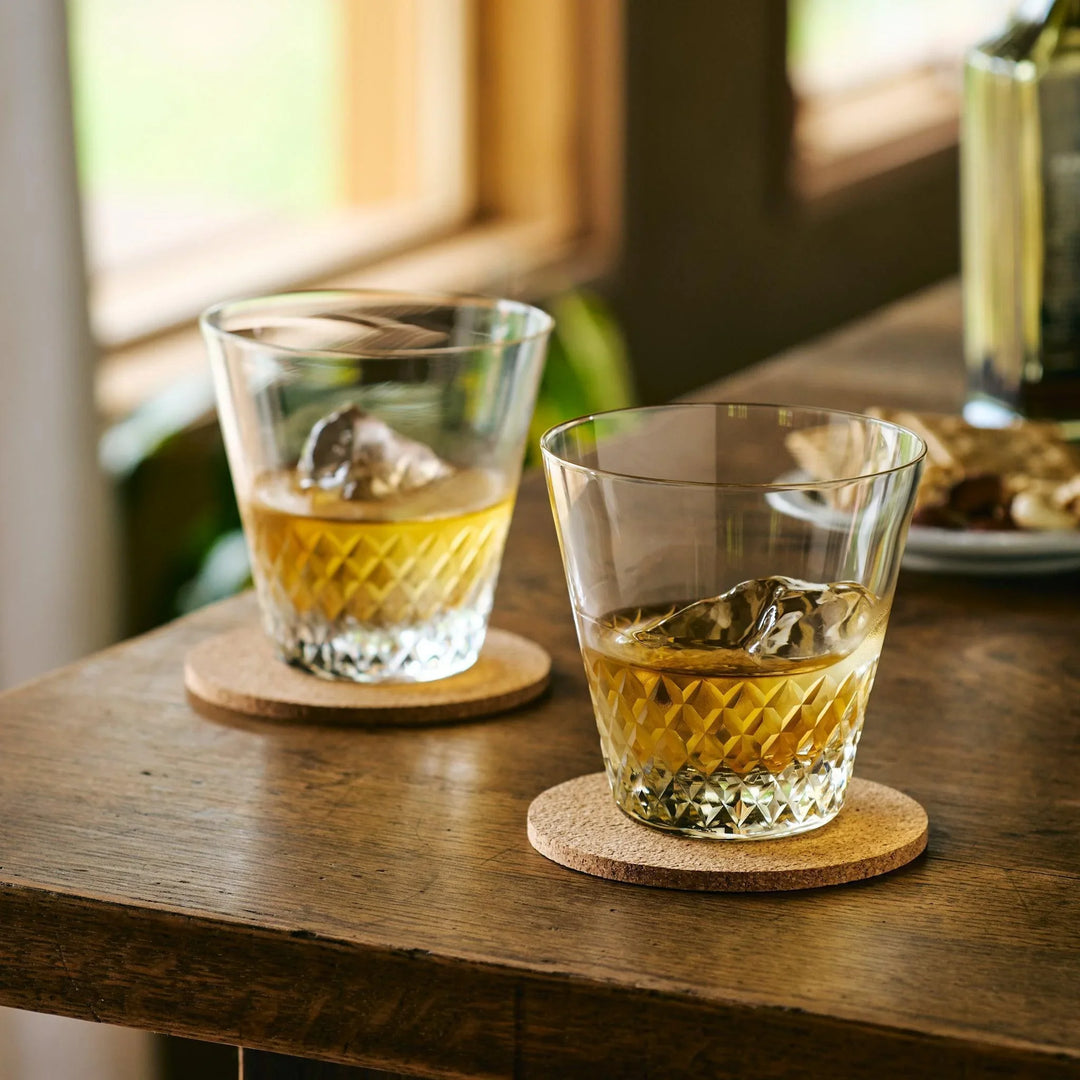 Two diamond-cut Japanese whiskey glasses with whisky and ice, placed on cork coasters on a wooden table.
