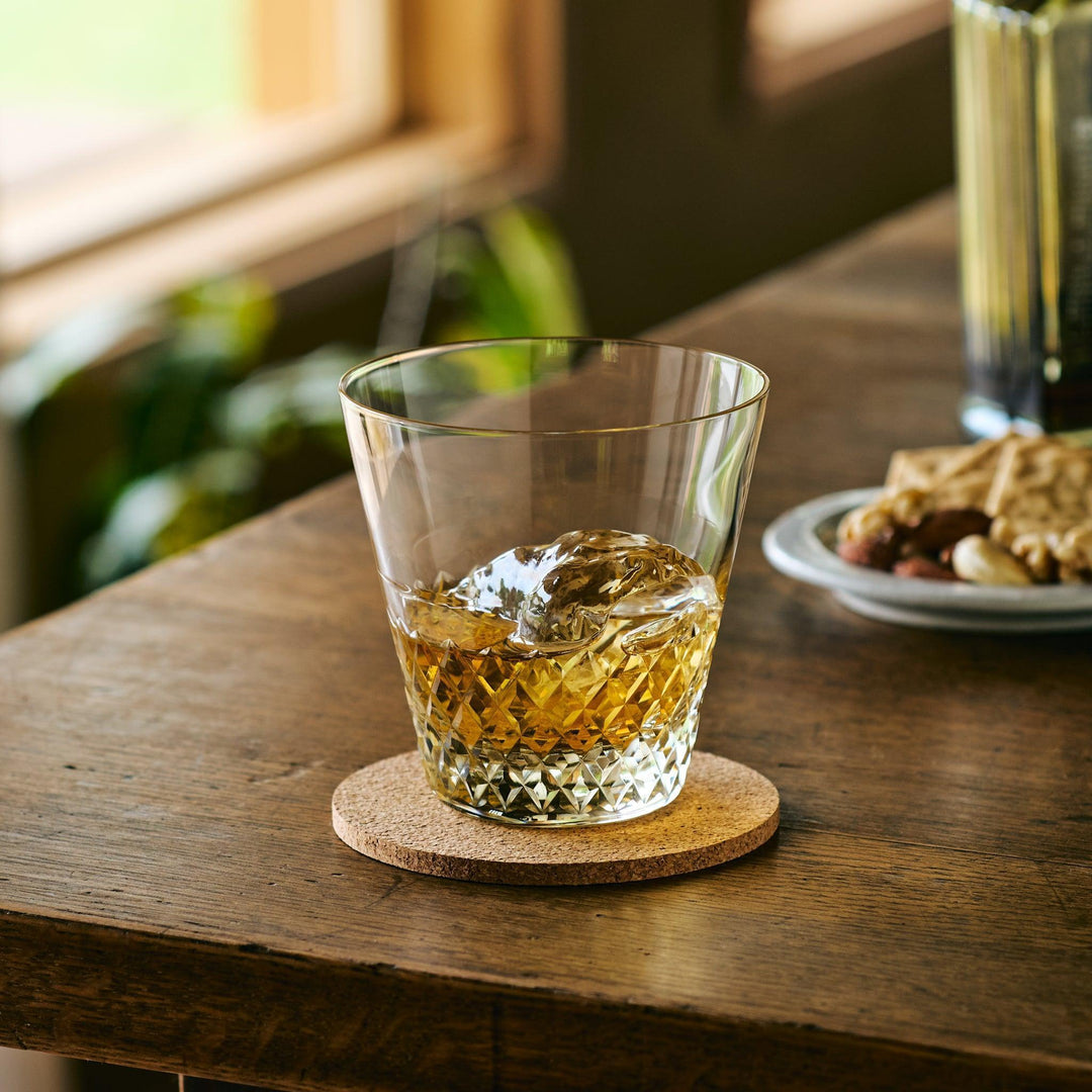 Faceted Japanese whiskey glass filled with whisky and a large ice cube, placed on a cork coaster.