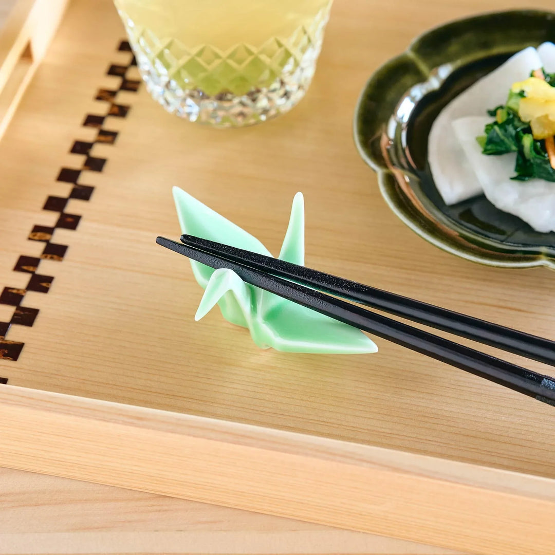 A green origami crane-shaped chopstick rest supporting black chopsticks, placed on a light wooden tray.