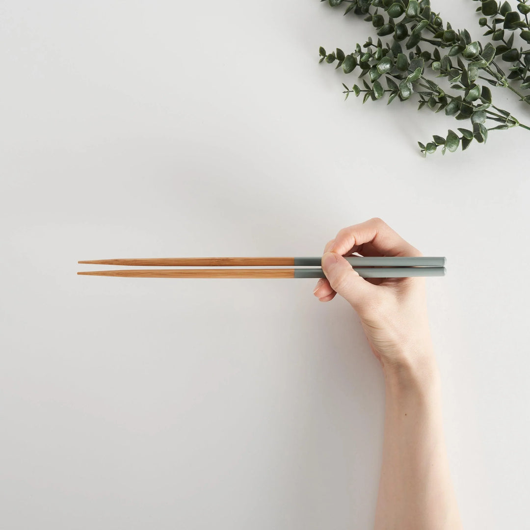 Hand holding light gray and bamboo chopsticks, showcasing the length and ergonomic design.