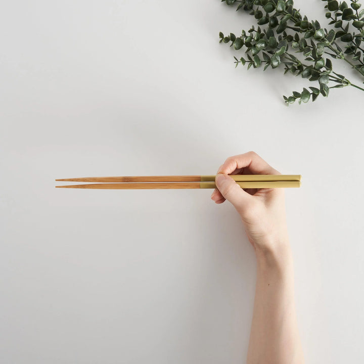 Hand holding yellow-tipped cooking chopsticks with wooden stems, with green foliage in the background.
