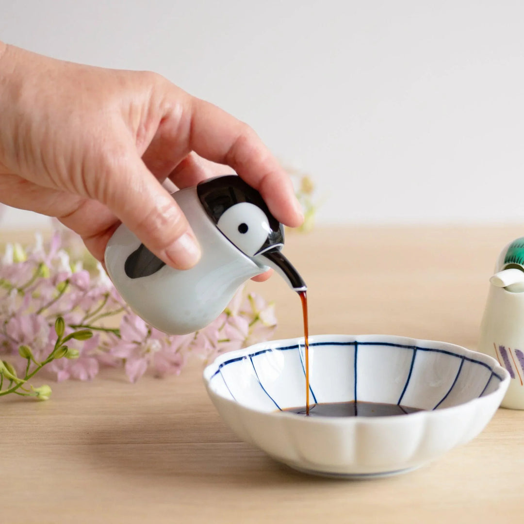 Penguin shaped soy sauce dispenser pouring into scalloped white dish with navy lines and pink flowers.