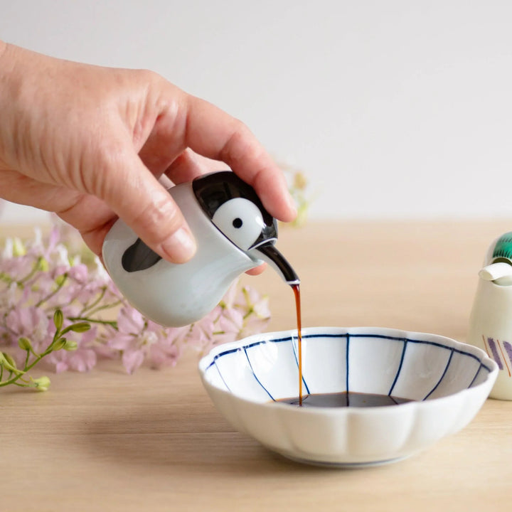 Penguin shaped soy sauce dispenser pouring into scalloped white dish with navy lines and pink flowers.