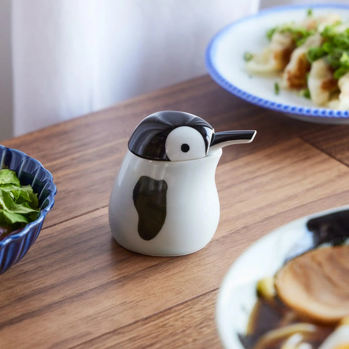 A ceramic soy sauce dispenser shaped like a cute penguin, placed on a wooden table alongside Japanese dishes.