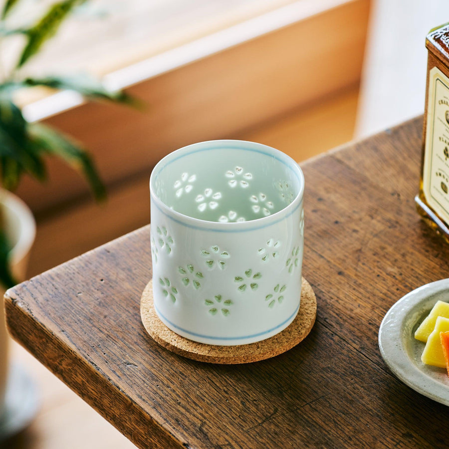 A white porcelain whiskey cup with delicate sakura cutout patterns, placed on a cork coaster by a wooden table.