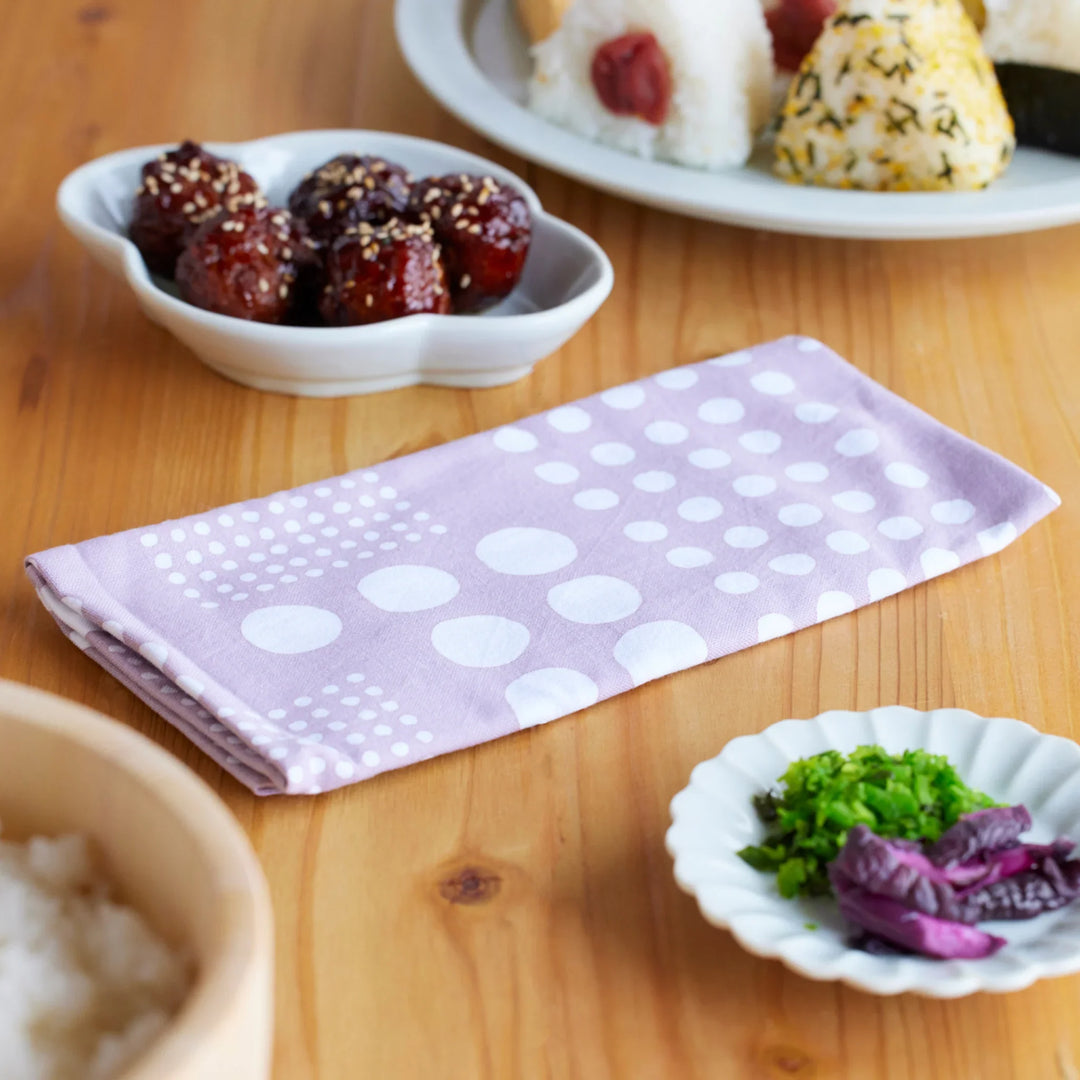 Purple polka dot tenugui towel on wooden table beside rice pickles and onigiri for homely scene.