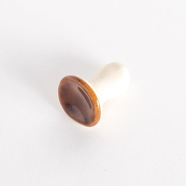 A porcelain chopsticks rest shaped like a porcini mushroom, with a glossy brown cap and smooth white stem.