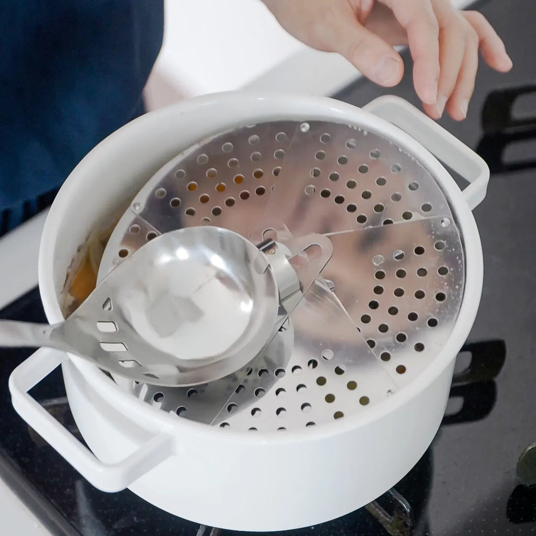 Stainless steel drop lid placed inside a white pot on the stove, with a slotted ladle resting on top for easy removal.