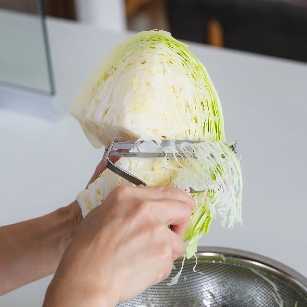 Nami using a vegetable peeler to shred green cabbage into a metal bowl.