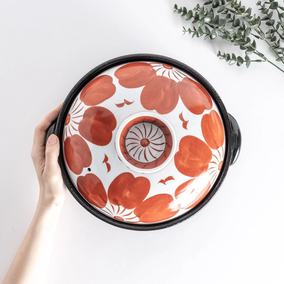 Hands lifting lid of Japanese donabe pot with red floral pattern on stove surrounded by small side dishes.