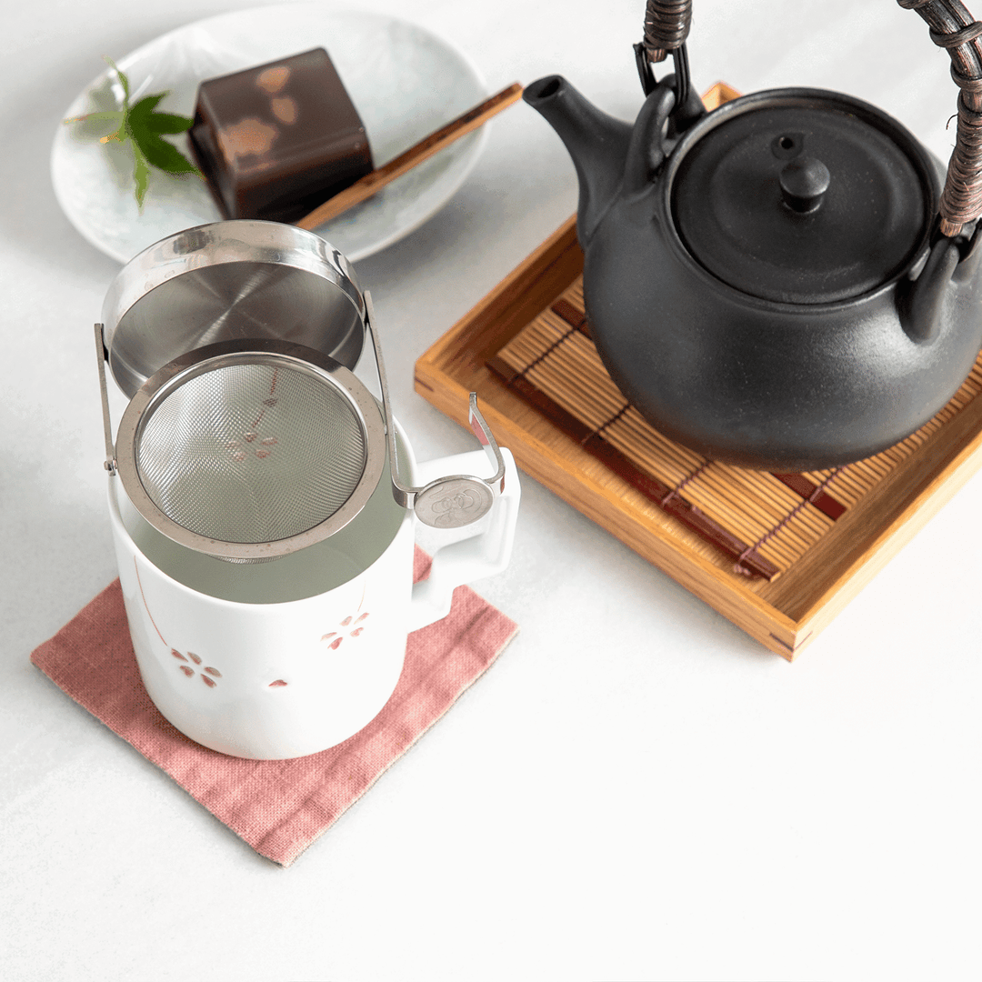 Japanese tea scene with a black teapot, rotating stainless tea strainer over a white mug, and yokan dessert on a plate