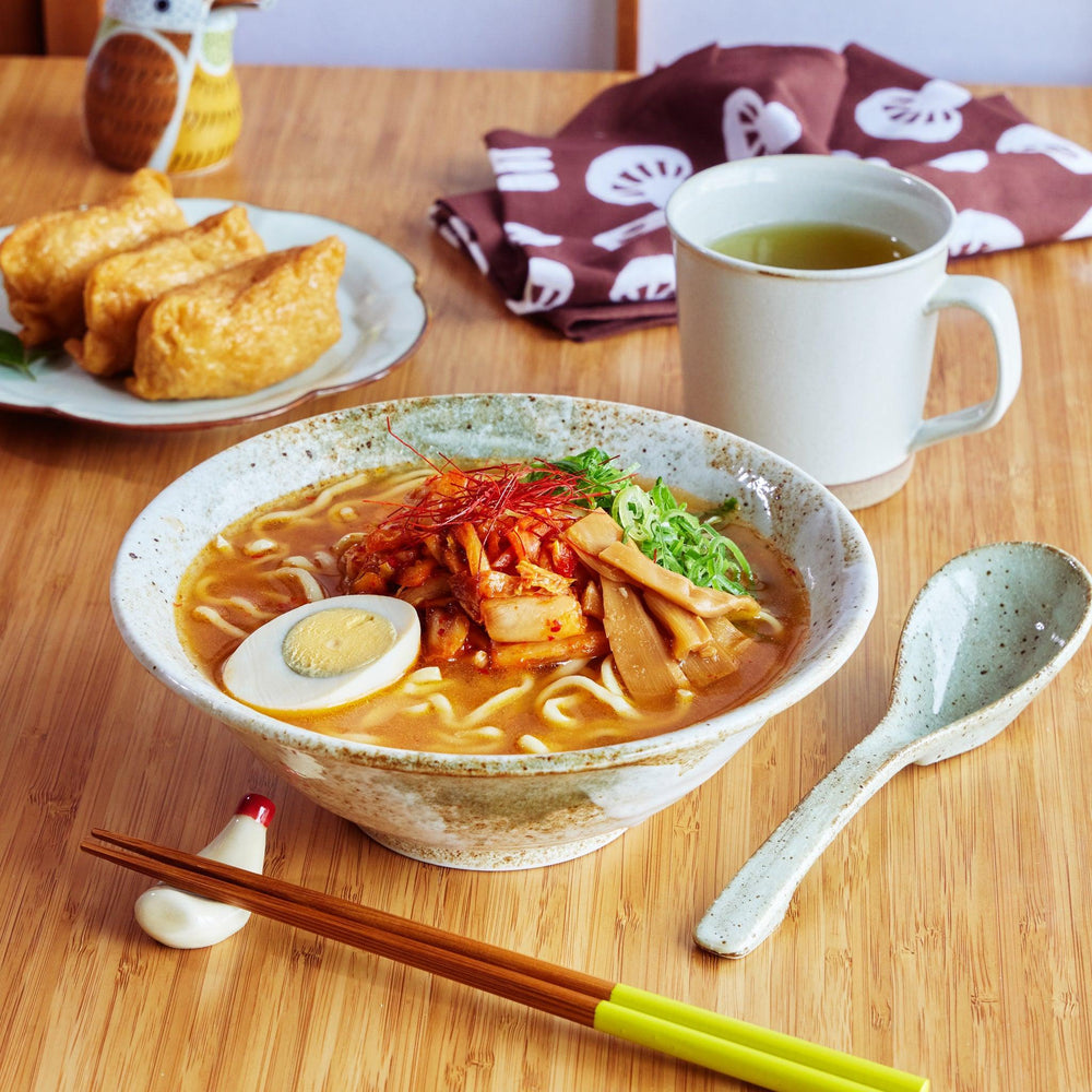 Handcrafted ceramic ramen bowl with spicy toppings and matching ceramic spoon, placed on a wooden table.