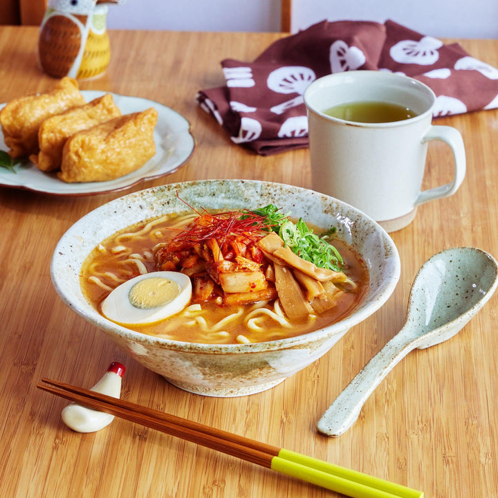 Handcrafted ceramic ramen bowl with spicy toppings and matching ceramic spoon, placed on a wooden table.