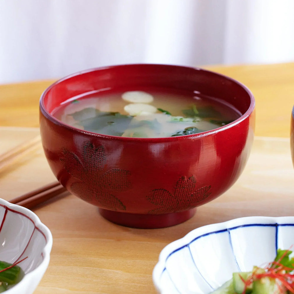 A red lacquered miso soup bowl with delicate sakura motifs, holding a warm miso soup with tofu and greens.