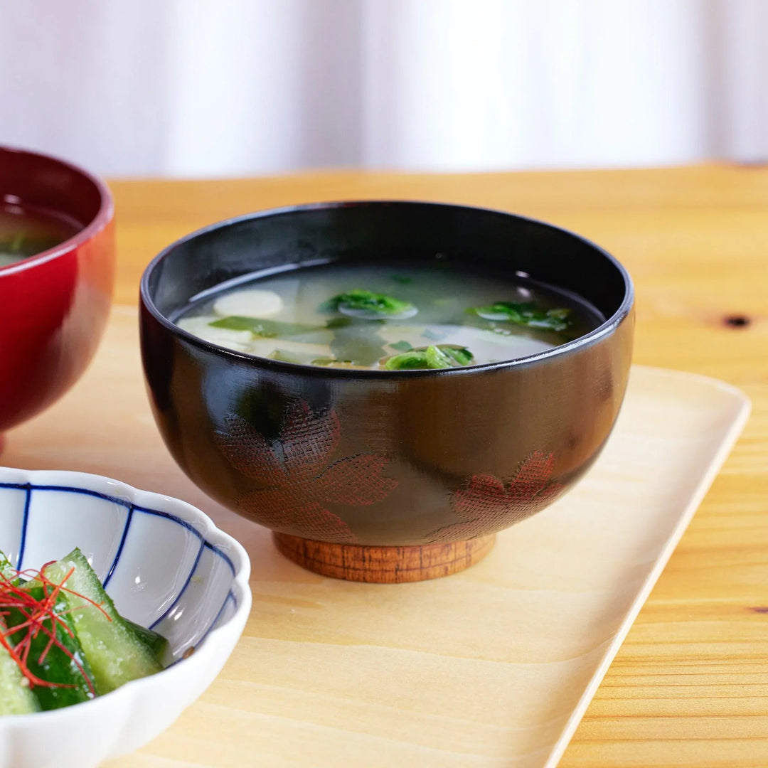 A black lacquered miso soup bowl with subtle sakura patterns, filled with clear broth and green vegetables.