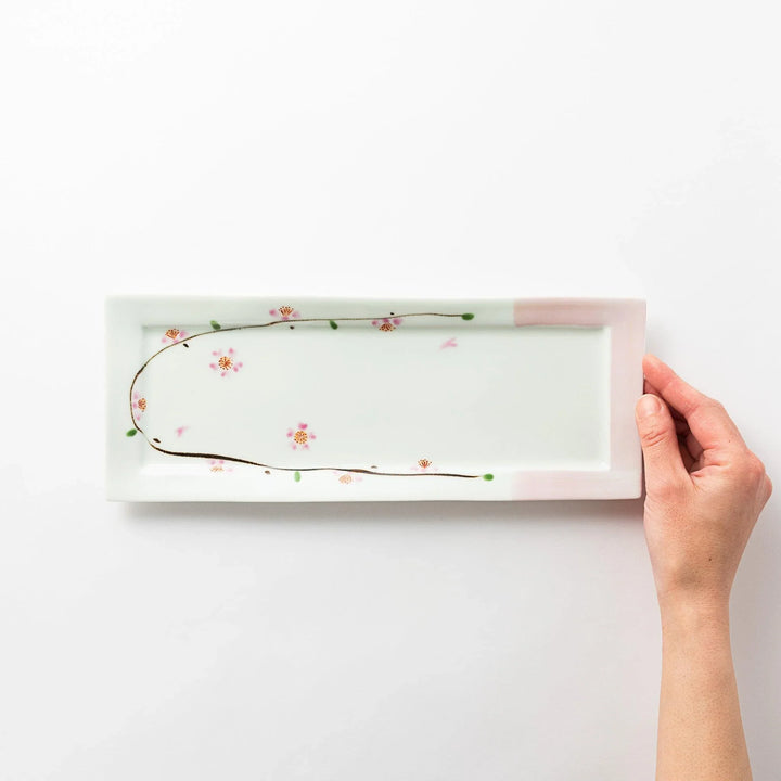 Hand holding a rectangular plate viewed from above, showcasing the delicate cherry blossom pattern with pink accents.