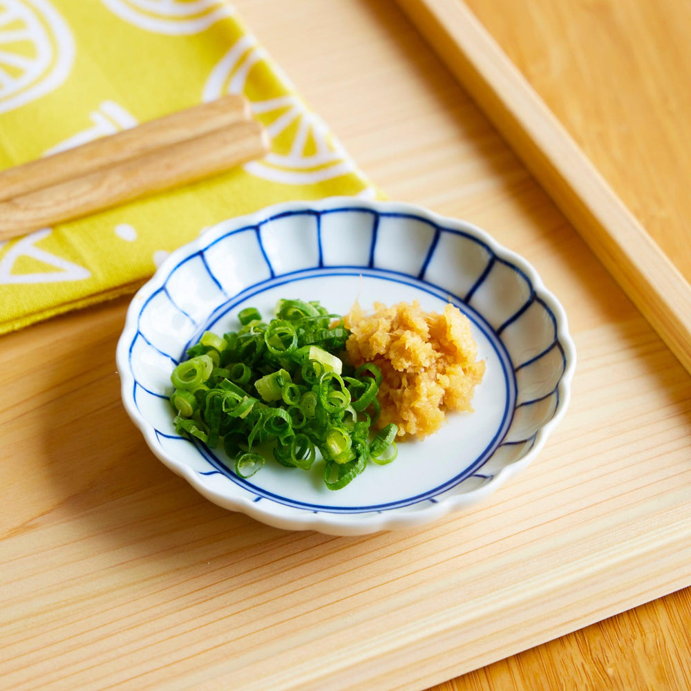 White scalloped sauce dish with navy lines holding chopped green onions and grated ginger on tray.
