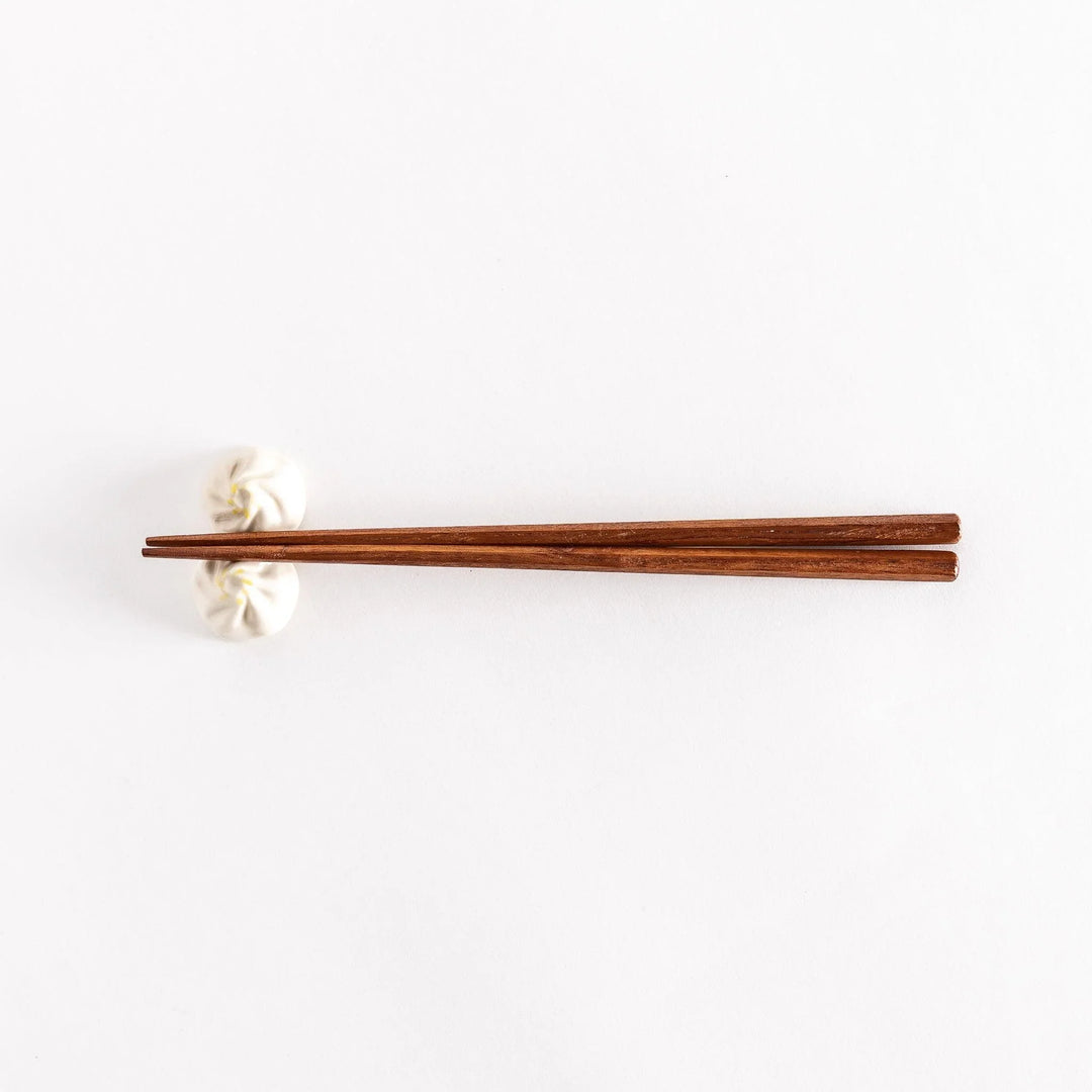 Wide shot of wooden chopsticks resting on a soup dumpling-shaped chopstick rest, placed on a white surface.