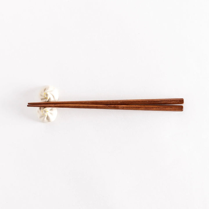 Wide shot of wooden chopsticks resting on a soup dumpling-shaped chopstick rest, placed on a white surface.