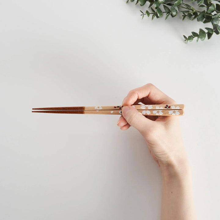 A hand gracefully holds chopsticks with a delicate floral design, featuring white flowers with a red accent.