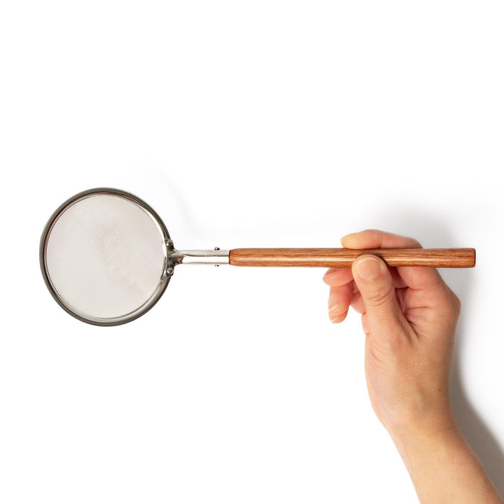 Stainless steel round-mesh skimmer with long wooden handle, perfect for straining soups and frying in home kitchens.