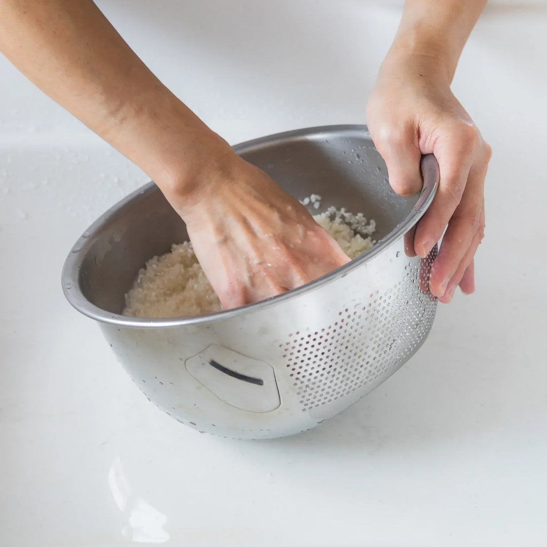 Nami washing rice by hand in a stainless steel colander, swirling the grains to clean them evenly.