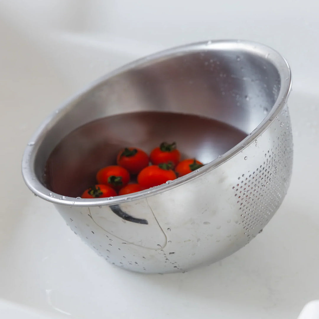 Cherry tomatoes soaking in water inside a stainless steel colander, ready to be washed in the kitchen sink.