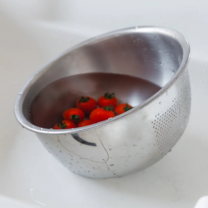 Cherry tomatoes soaking in water inside a stainless steel colander, ready to be washed in the kitchen sink.