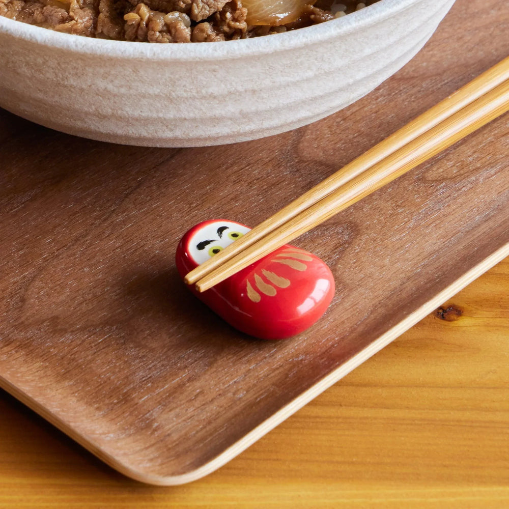 A red ceramic chopstick rest shaped like a Daruma doll with gold stripes, holding a pair of bamboo chopsticks.
