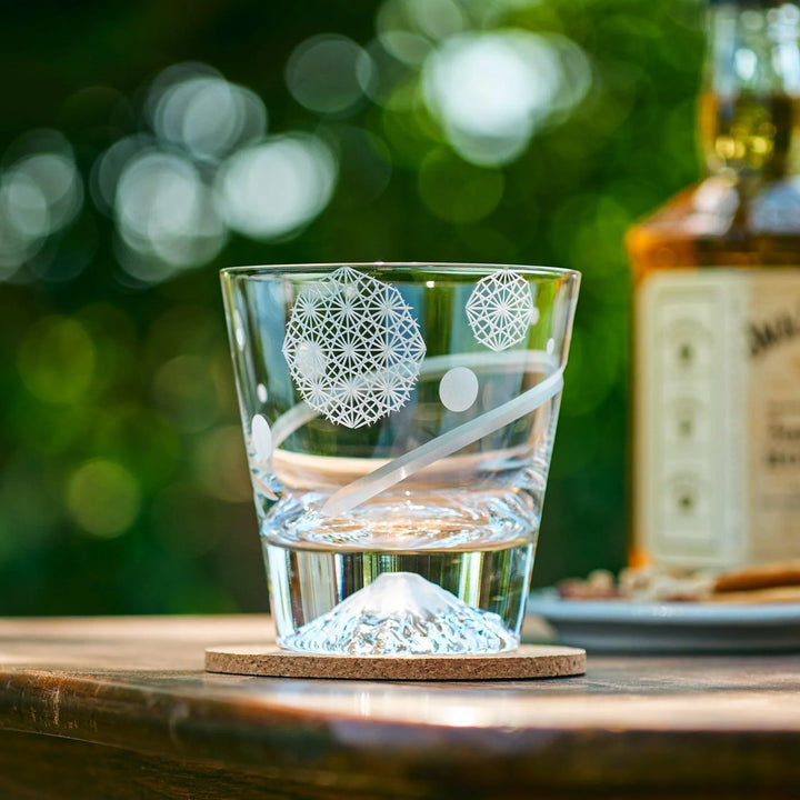 A clear Japanese glass with etched Asanoha patterns and Mt. Fuji-shaped base, placed on a wooden table in sunlight.