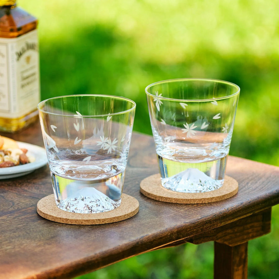 Clear glasses with sakura engravings and Mt. Fuji-shaped bottoms, placed on cork coasters outdoors.