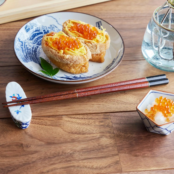 A pair of vermilion and black urushi lacquered chopsticks with fine line detailing, placed beside a plate of sushi.