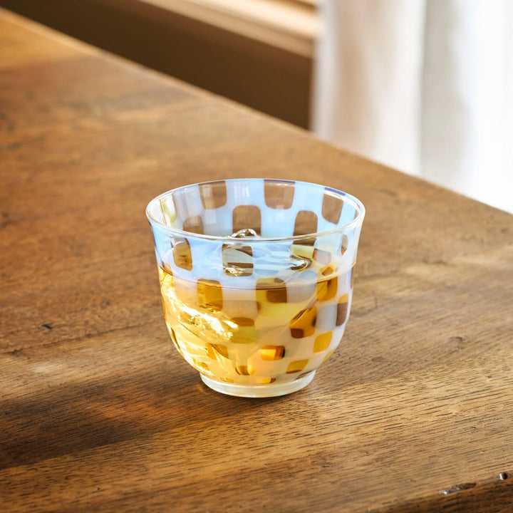 A vintage glass tea cup featuring a light blue checkerboard design, filled with amber tea, set on a wooden table.
