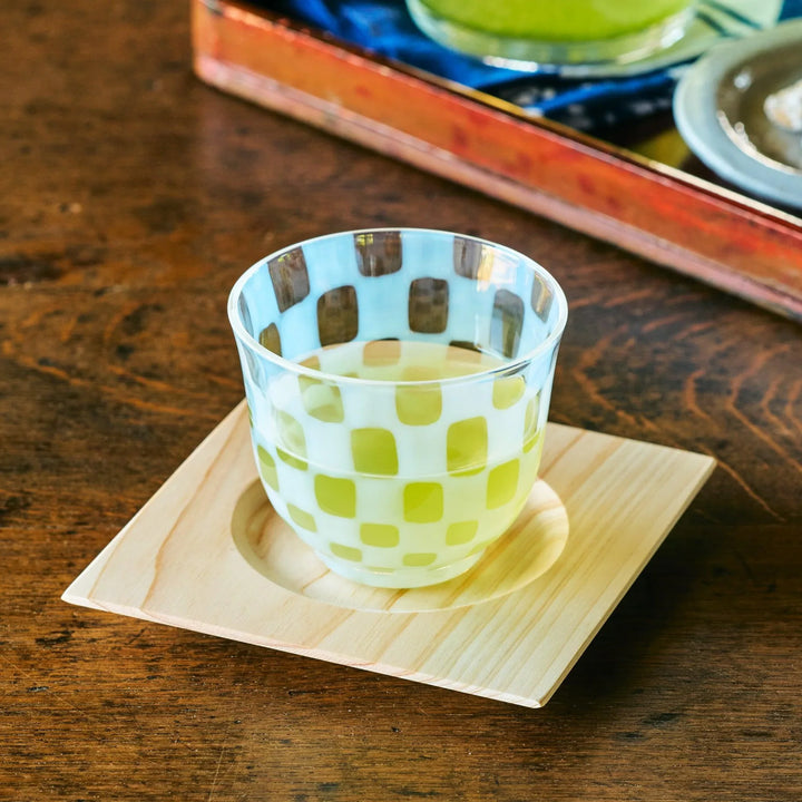 A Japanese glass tea cup with a translucent checkered pattern, filled with green tea and set on a square wooden coaster.
