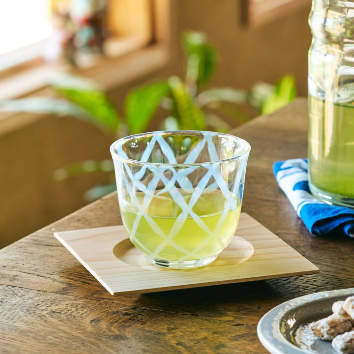 A glass tea cup with a white lattice pattern, filled with green tea and placed on a wooden coaster indoors.