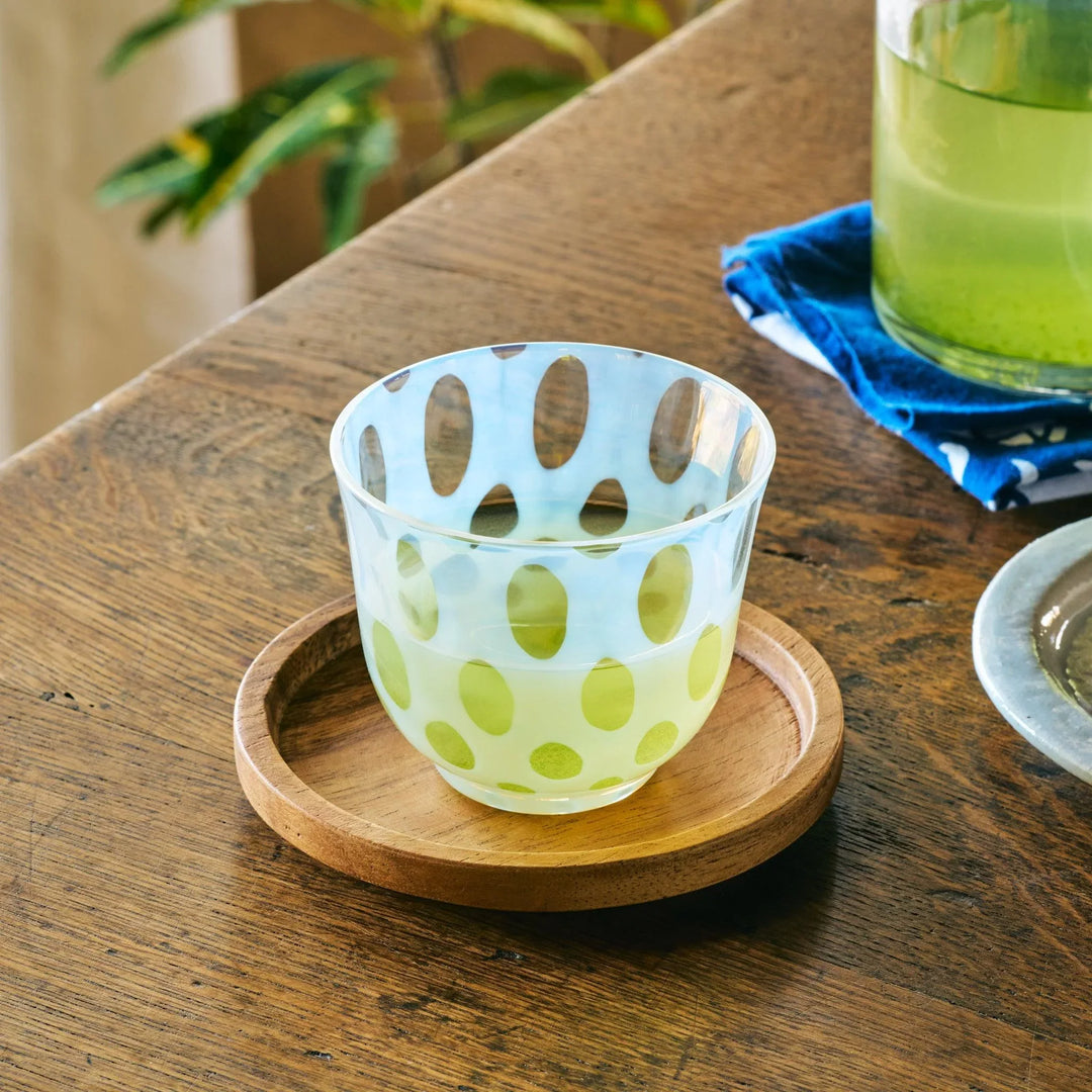 A glass tea cup with oval dot patterns, filled with green tea and placed on a round wooden coaster indoors.