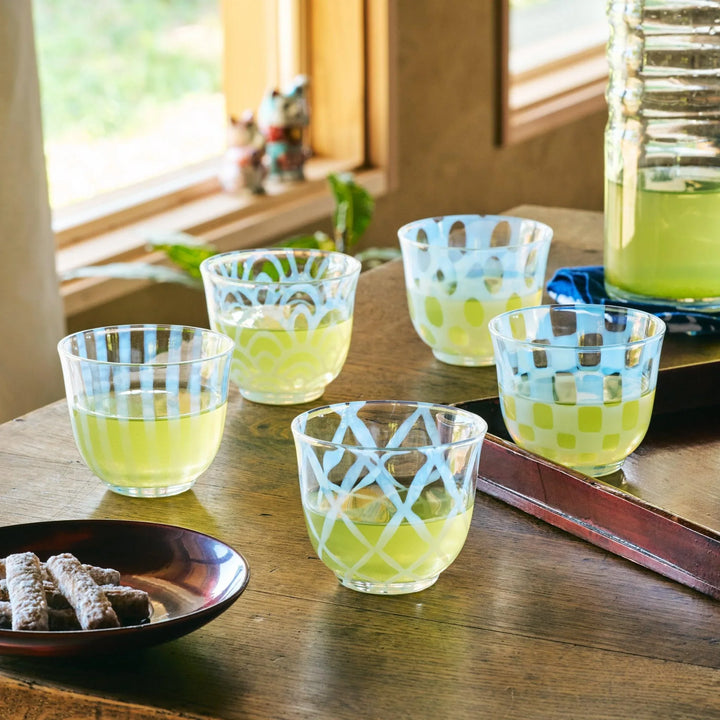 Five patterned glass tea cups filled with green tea, each featuring a unique blue design, set on a wooden table.