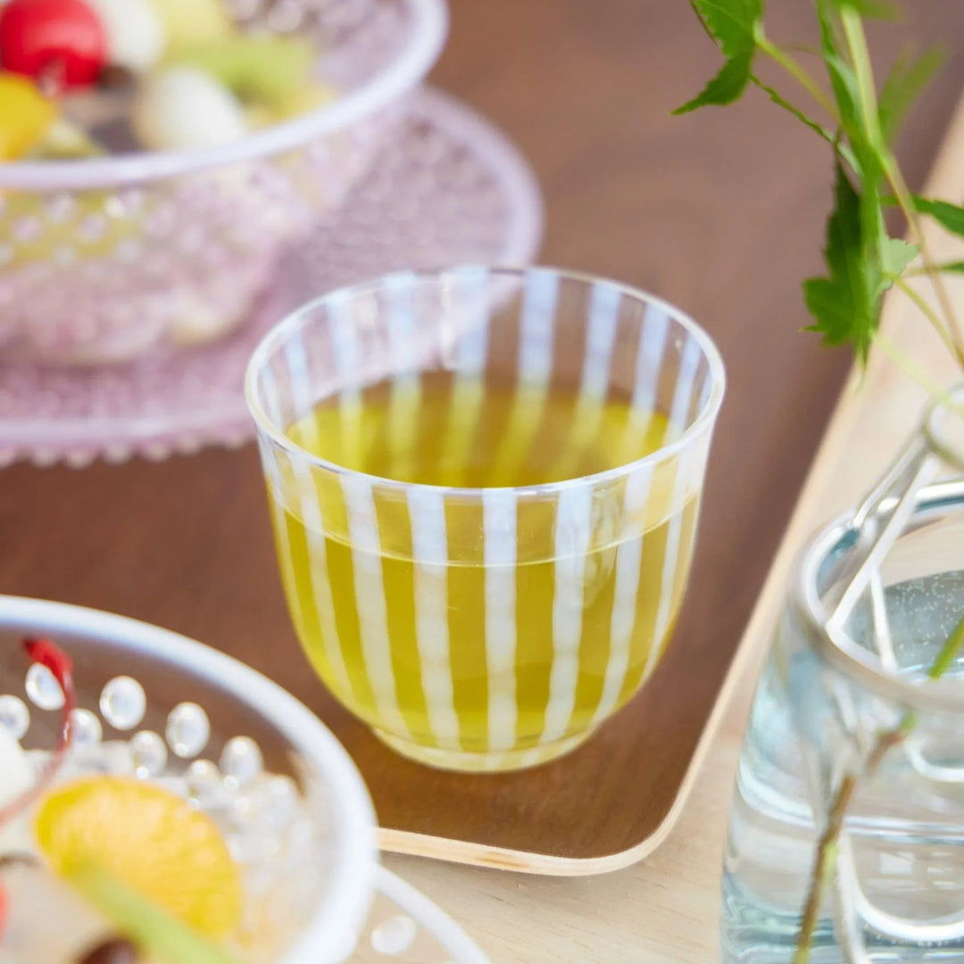 Clear glass tea cup with vertical blue stripes, filled with green tea and placed on a wooden tray with summer tableware.