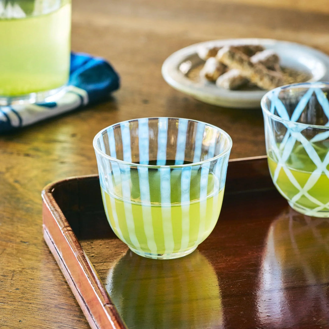 A glass tea cup with vertical blue stripes, filled with green tea and placed on a wooden tray in warm light.