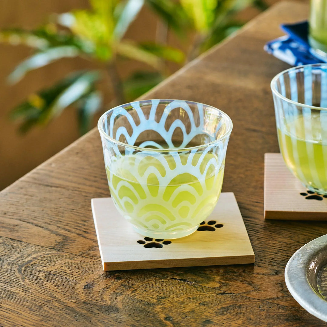 A glass tea cup with Seigaiha wave patterns and green tea, placed on a wooden coaster with a paw print cutout.