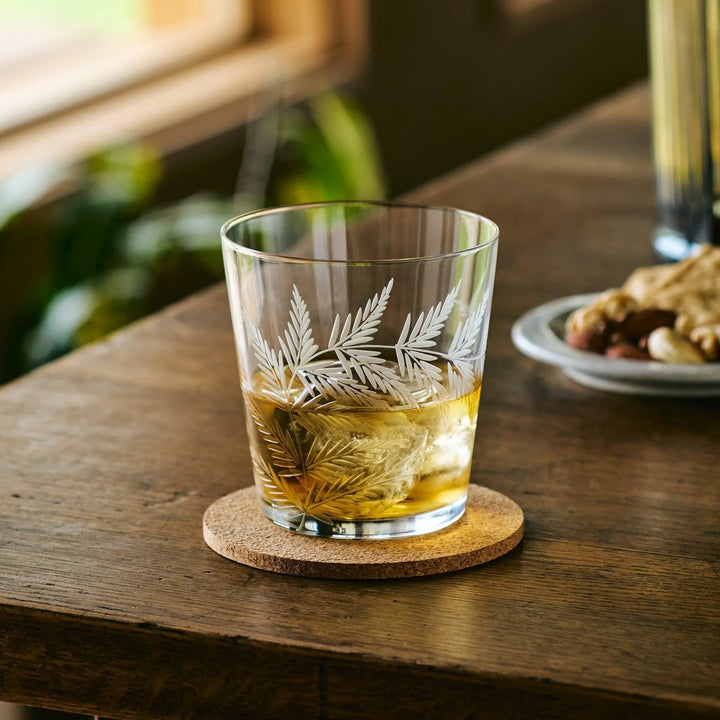 Japanese whiskey glass with etched leaf design, filled with whisky and ice, on a cork coaster on a wooden table.