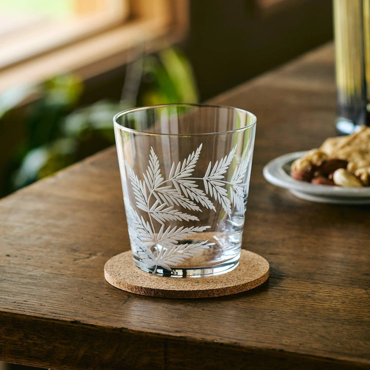 Empty Japanese whiskey glass with white etched leaf pattern, placed on a cork coaster on a wooden table.