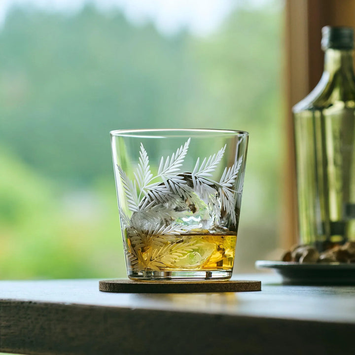 Etched Japanese whiskey glass with a leaf design, filled with whisky and ice, placed on a cork coaster.