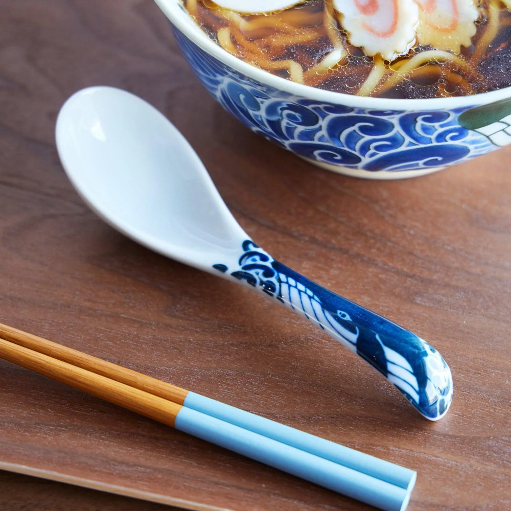 A close-up of a ceramic ramen spoon with a whale motif on the handle, placed beside blue chopsticks and a ramen bowl.