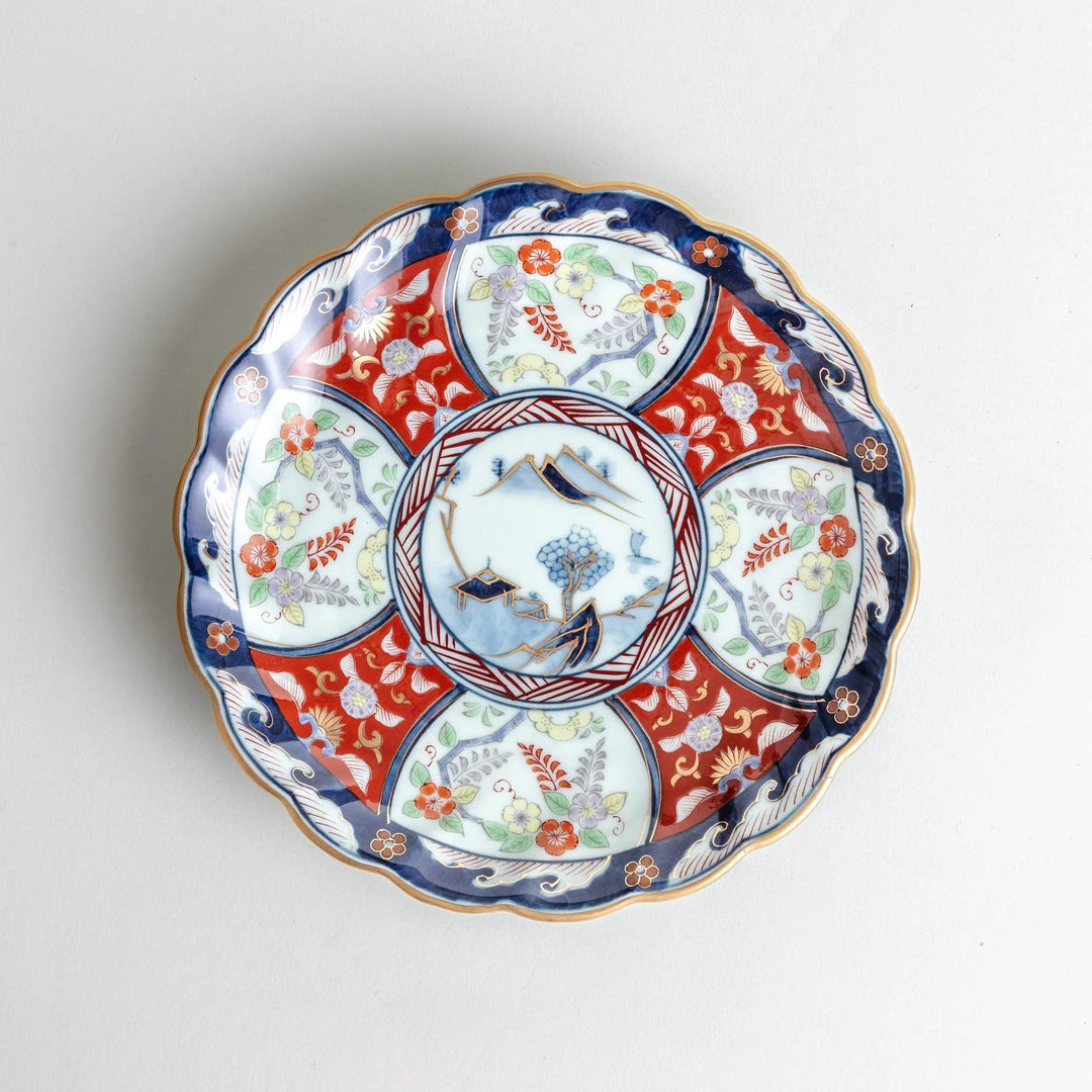 Scalloped porcelain bowl with Imari style red and blue alternating patterns and central landscape motif.