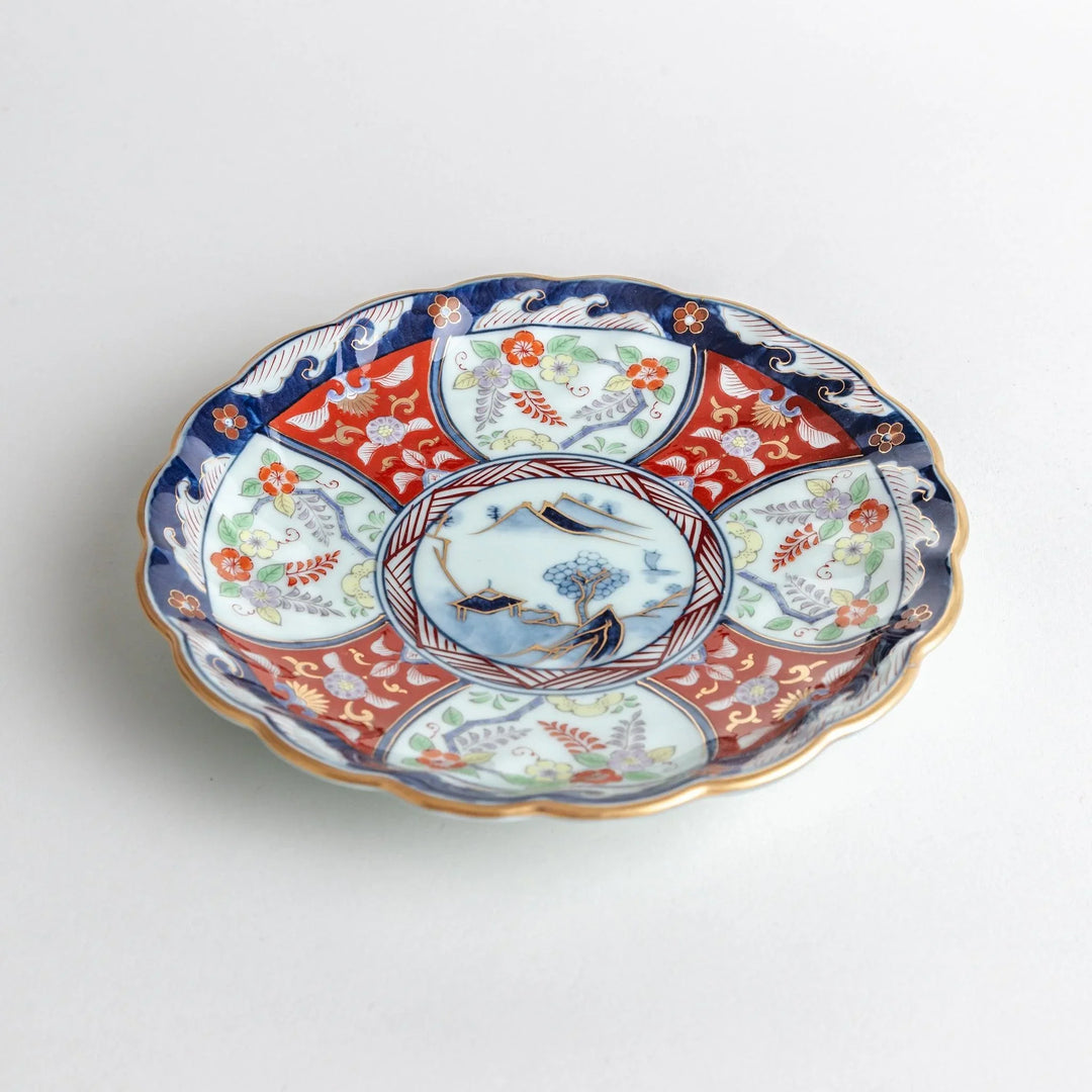 Scalloped porcelain bowl with Imari style red and blue alternating patterns and central landscape motif.