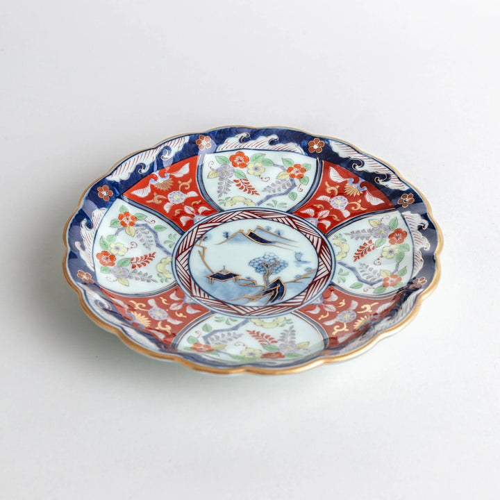 Scalloped porcelain bowl with Imari style red and blue alternating patterns and central landscape motif.